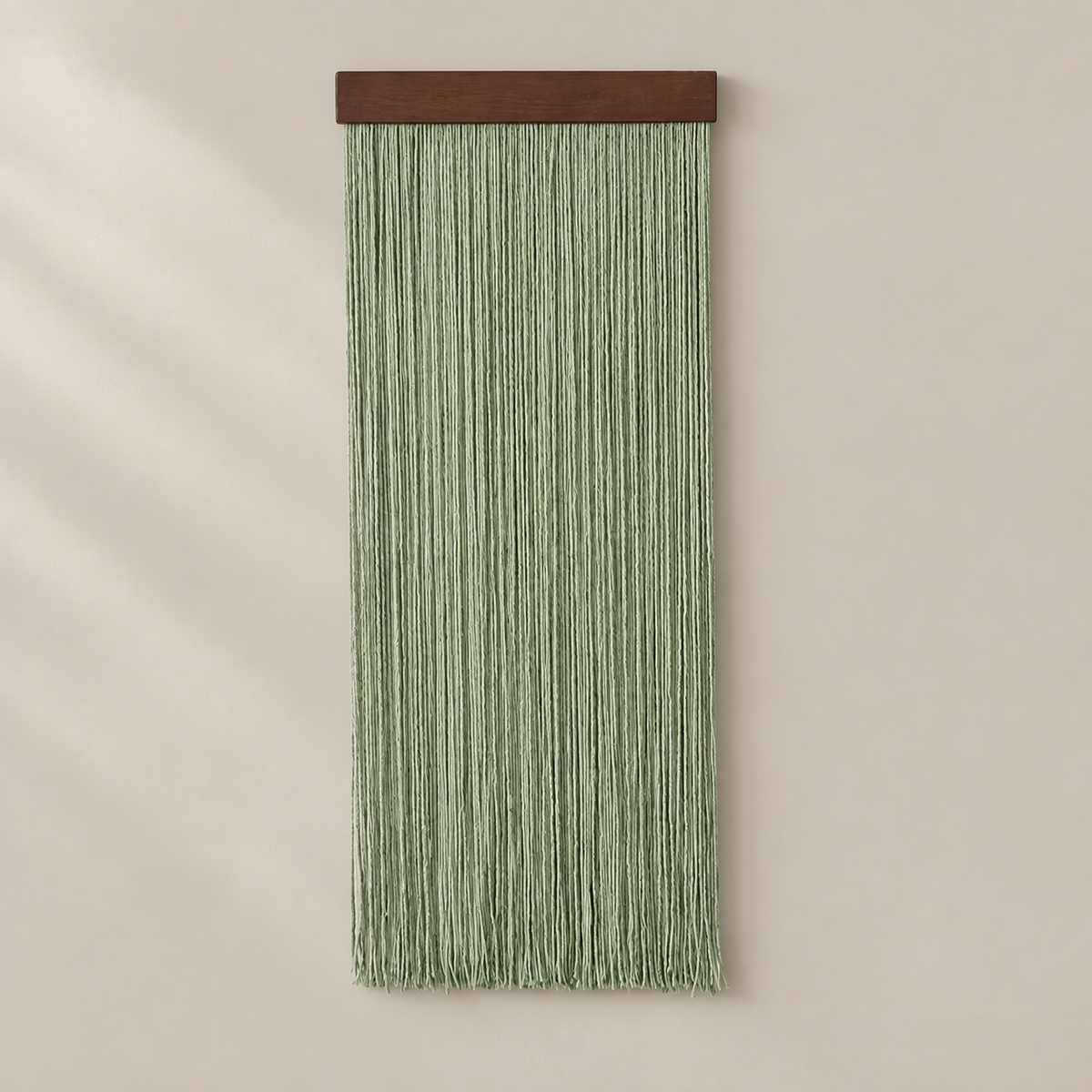 A vertical sage green macrame wall hanging with a dark brown wooden top bar, displayed against a warm beige wall with soft diagonal shadows.