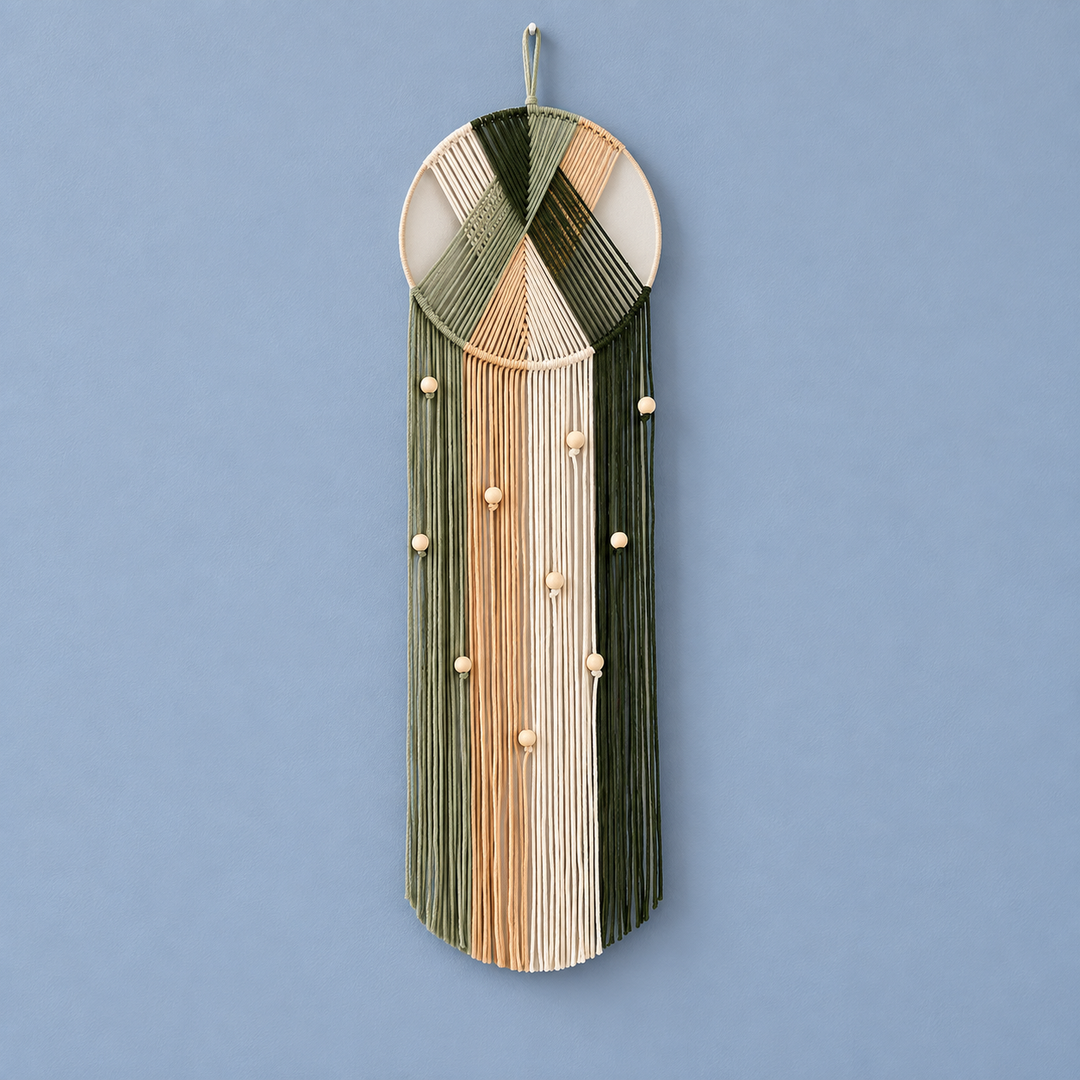A bohemian-style woven wall hanging with a circular frame, vertical fringe strands in olive green, beige, and tan, and small wooden beads, shown against a solid light blue background.