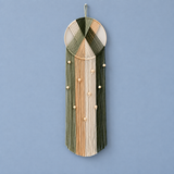 A bohemian-style woven wall hanging with a circular frame, vertical fringe strands in olive green, beige, and tan, and small wooden beads, shown against a solid light blue background.
