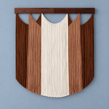 A bohemian-style wall hanging macrame wall decor featuring vertical fringed panels in shades of brown and cream, suspended from a dark wooden bar against a solid blue-gray wall.