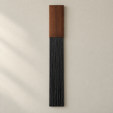 A modern wall-mounted decorative piece featuring a rectangular medium-brown wood top and a long, narrow black fringed or corded lower section, shown against a warm off-white wall.