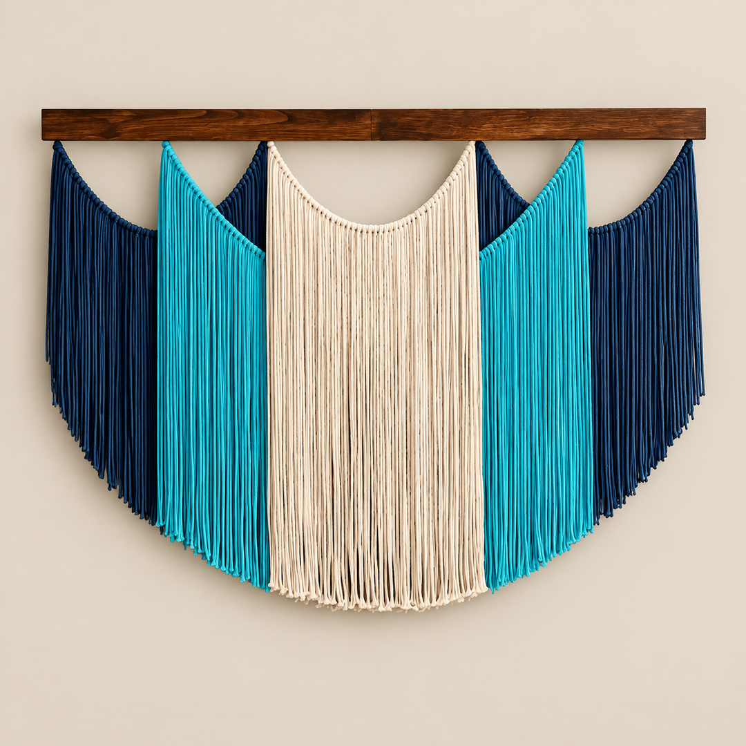 A bohemian-style macrame wall hanging with a semi-circular design, featuring a central cream-colored fringed panel flanked by turquoise and dark navy blue panels, suspended from a dark brown wooden bar against a light beige wall.