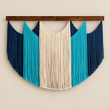 A bohemian-style macrame wall hanging with a semi-circular design, featuring a central cream-colored fringed panel flanked by turquoise and dark navy blue panels, suspended from a dark brown wooden bar against a light beige wall.
