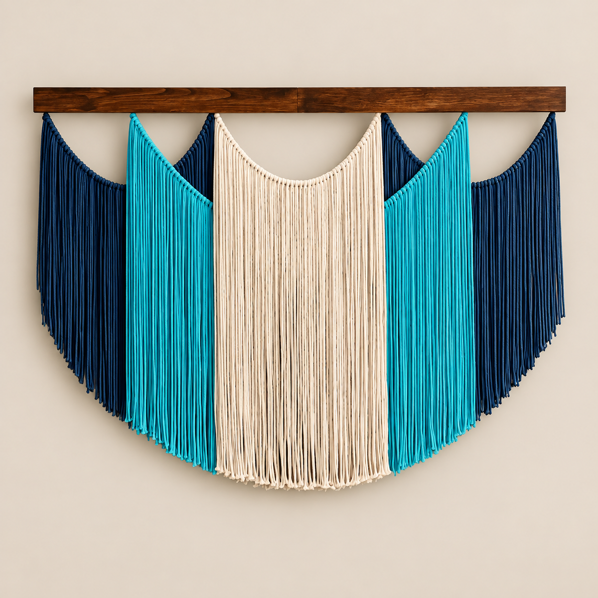 A bohemian-style macrame wall hanging with a semi-circular design, featuring a central cream-colored fringed panel flanked by turquoise and dark navy blue panels, suspended from a dark brown wooden bar against a light beige wall.