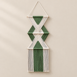 A geometric macrame wall hanging with green and off-white woven cords, suspended from a wooden dowel against a warm beige wall.