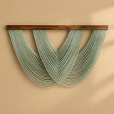 A wall-mounted decorative sculpture made of many thin pale green hanging strands suspended from a dark brown wooden bar, set against a warm beige wall.