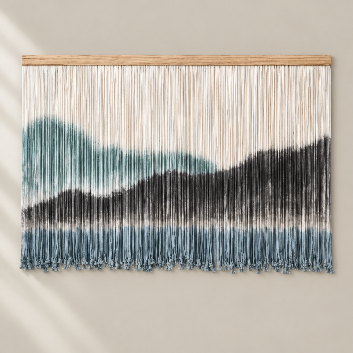 A horizontal wall hanging with a wooden top bar and many hanging fringe strands, featuring an abstract mountain-like pattern in off-white, teal, and charcoal gray on a warm beige wall.