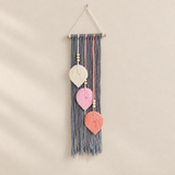 A bohemian-style macrame wall hanging with a gray tassel fringe, decorated with three hand-stitched leaf shapes in cream, pink, and coral, suspended from a wooden dowel with a hanging string.