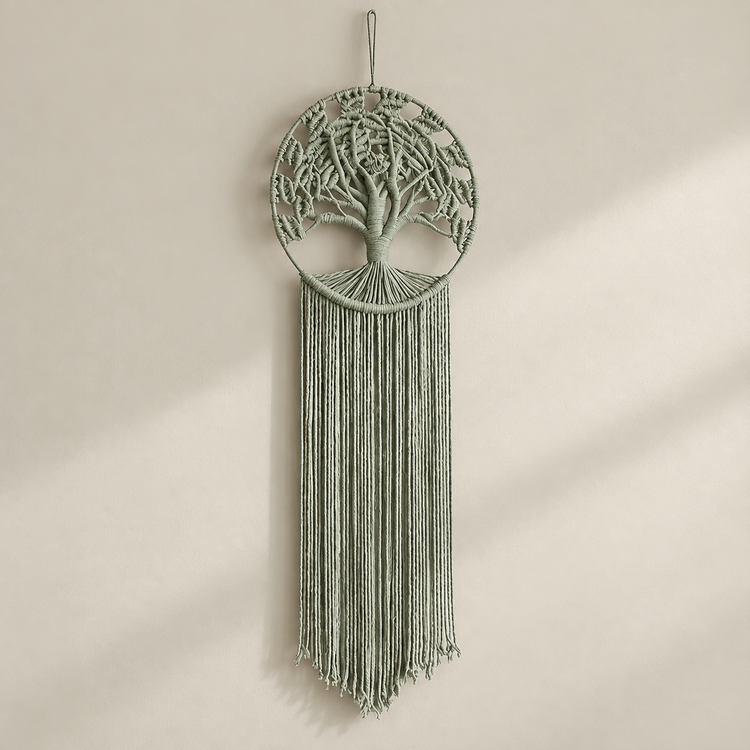 A sage green macrame wall hanging featuring a tree of life design inside a circular hoop with long fringe, suspended against a warm beige wall with soft diagonal shadows.