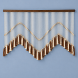 A bohemian-style wall hanging macrame wall decor with a dark wood top bar, cream and brown cord strands, and tassel accents, displayed against a solid light blue wall.