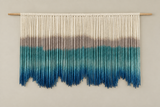 A handmade macrame wall hanging suspended from a light wood dowel, featuring a cream-colored top section that fades into taupe and teal, ending in vibrant blue tassels at the bottom, set against a plain beige wall.