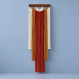 A vertical woven wall hanging with layered fringe strands in cream, mustard, rust red, and terracotta, suspended from a dark wood bar against a muted blue-gray wall.
