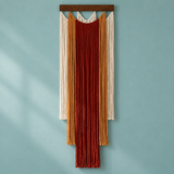 A vertical boho macrame wall hanging with layered fringe strands in cream, mustard, rust red, and dark brown, suspended from a dark wood bar against a muted blue wall.