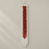 A vertical macrame wall hanging with a red woven central panel and long white fringe, suspended from a light wood dowel against a plain warm gray wall.