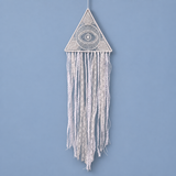 A white macrame wall hanging with a triangular top featuring an eye motif and long fringe strands, shown against a solid light blue background.