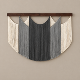 A bohemian-style macrame wall hanging with a dark wood top bar, featuring vertical fringe in cream, light gray, and charcoal gray, suspended against a solid beige wall.