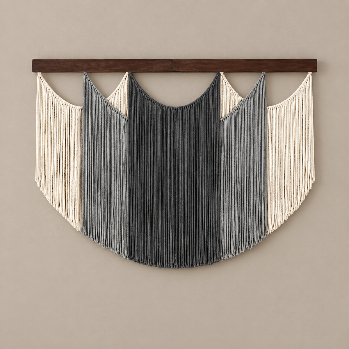 A bohemian-style macrame wall hanging with a dark wood top bar, featuring vertical fringe in cream, light gray, and charcoal gray, suspended against a solid beige wall.