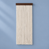 A vertical macrame wall hanging with cream-colored cord and a dark brown wooden top bar, displayed against a light blue wall.