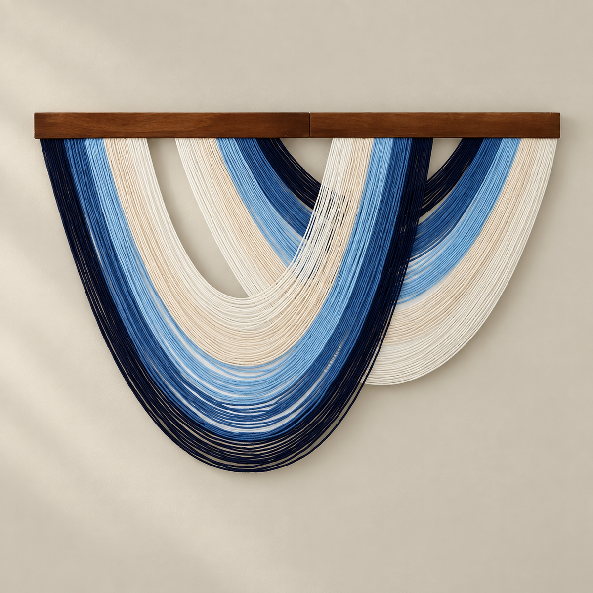A modern wall hanging sculpture made of layered, draped strands of dark navy, medium blue, light blue, cream, and white yarn suspended from a horizontal wooden bar, set against a warm beige wall.