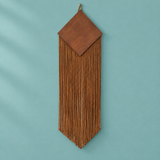 A brown leather wall hanging with a geometric diamond-shaped top and long fringe, suspended against a solid teal background.