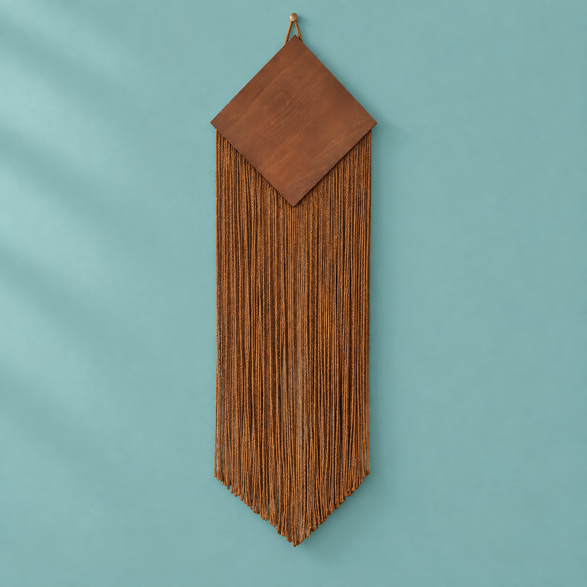 A brown leather wall hanging with a geometric diamond-shaped top and long fringe, suspended against a solid teal background.