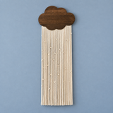 A cloud-shaped wall hanging with a dark brown wood top and cream-colored string fringe, shown against a solid blue background.