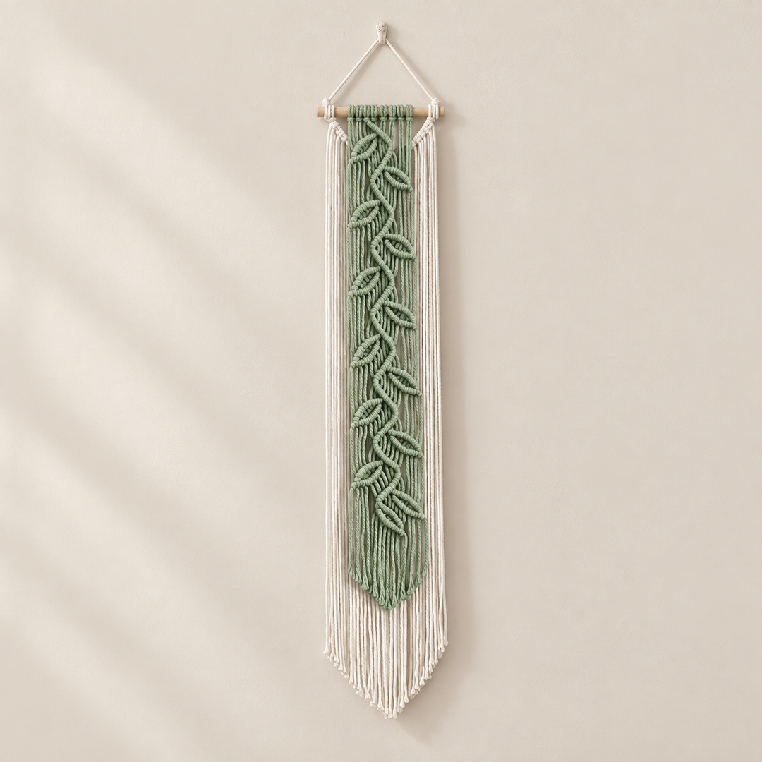 A vertical macrame wall hanging with a sage green leaf pattern in the center and white fringe, suspended from a light wood dowel against a warm beige wall.