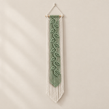 A vertical macrame wall hanging with a sage green leaf pattern in the center and white fringe, suspended from a light wood dowel against a warm beige wall.