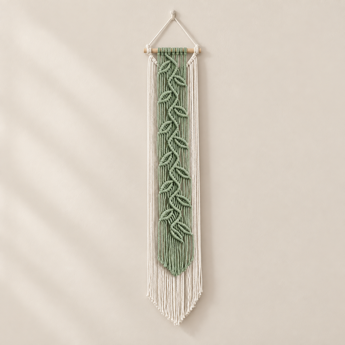 A vertical macrame wall hanging with a sage green leaf pattern in the center and white fringe, suspended from a light wood dowel against a warm beige wall.