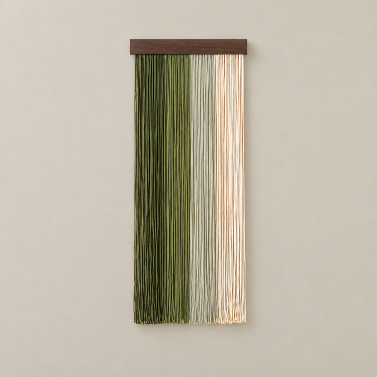 A vertical wall hanging with three narrow hanging strands in olive green, pale sage green, and cream, suspended from a dark brown wooden bar against a warm beige wall.