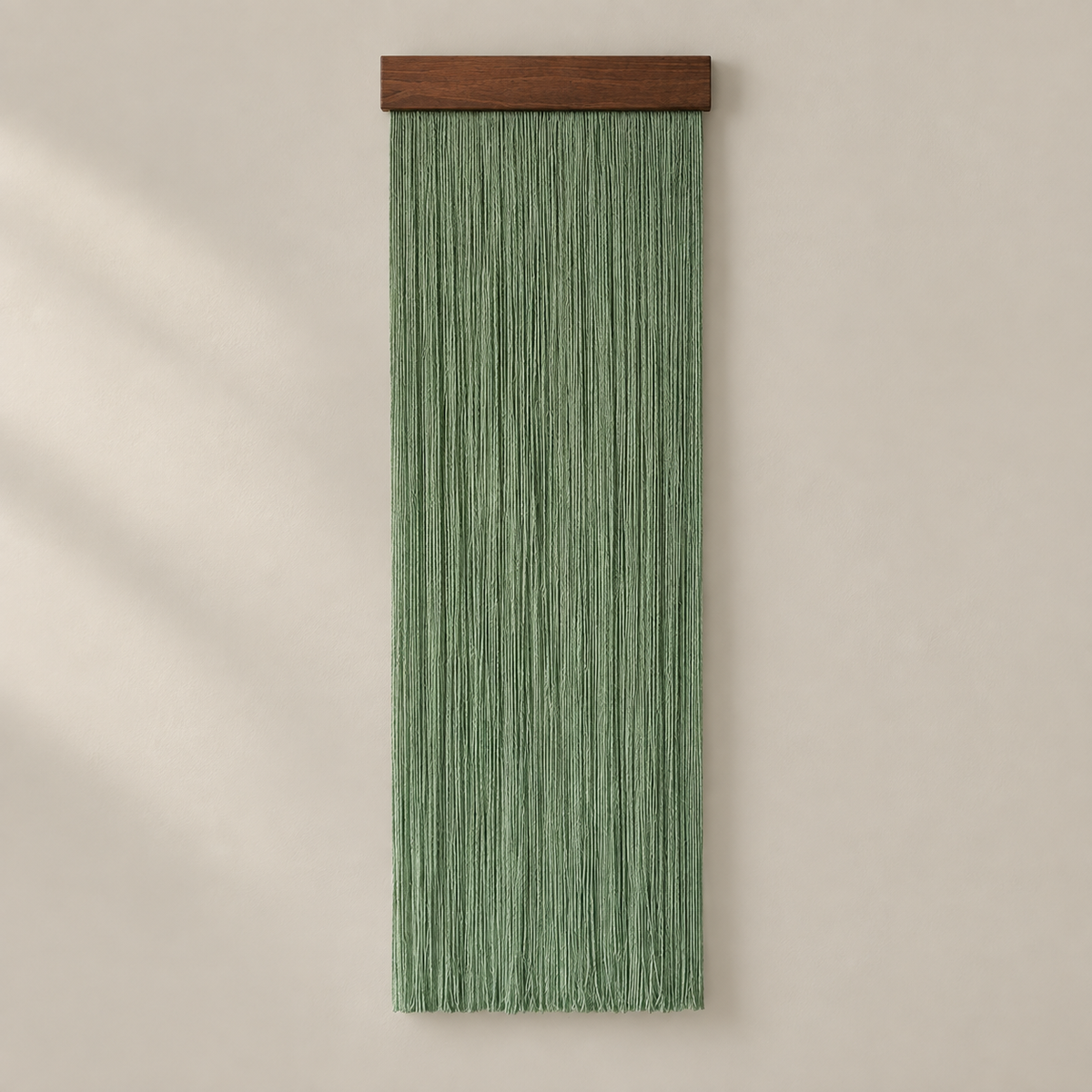 A vertical sage green macrame wall hanging with a dark brown wooden top bar, displayed against a warm beige wall with soft diagonal shadows.