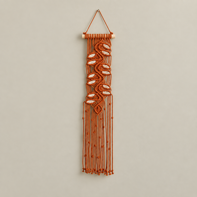 An orange macrame wall hanging with woven cowrie shells and a wooden dowel, suspended against a neutral beige wall.