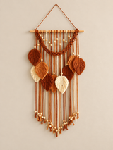 A bohemian-style woven wall hanging featuring a wooden dowel, braided cord, and multiple textured leaf shapes in warm brown, tan, and cream tones, suspended against a plain light beige wall.