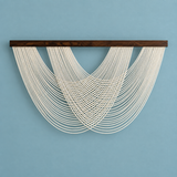 A wall-mounted macrame wall hanging with multiple cream-colored cords draped in a fan-like pattern from a dark brown wooden bar, set against a solid light blue background.