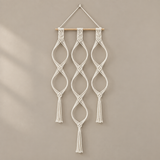 A bohemian-style macrame wall hanging with intricate white woven knots, three vertical hanging loops, and tassels, suspended from a light natural wood dowel on a neutral beige wall.