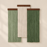 Three vertical woven wall hangings with long fringe, displayed against a warm off-white wall. The center hanging is cream, flanked by two sage green hangings, each with a dark brown wooden top bar.