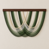 A wall-mounted decorative wall hanging with a dark wood top bar, featuring layered strands of dark and medium green thread with cream-colored beaded chains draped in a fan shape against a plain beige wall.
