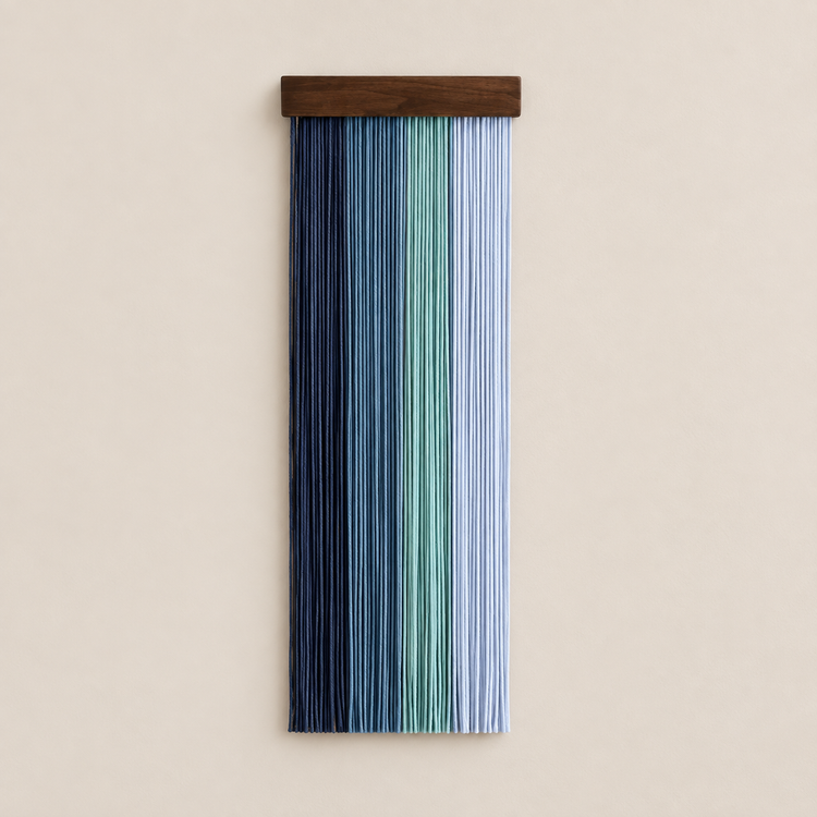 A vertical wall hanging with a dark wood top bar and multiple narrow hanging cords in vertical stripes of navy, blue, mint green, and pale blue, shown against a plain beige wall.