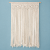 An off-white macrame wall hanging with a geometric woven pattern at the top and long fringe hanging below, suspended from a light natural wood dowel against a solid light blue wall.