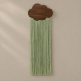 A decorative wall hanging with a dark brown wooden cloud-shaped top and long, narrow light sage green fringe strands hanging below, shown against a plain warm beige wall.