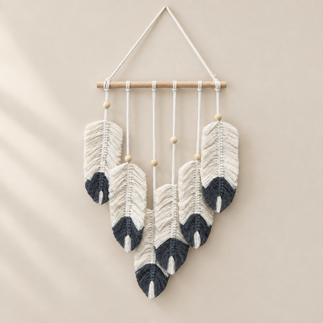 A bohemian-style woven wall hanging featuring five handwoven feathers in cream and charcoal gray, suspended from a light natural wood dowel with small wooden beads, set against a warm beige background.