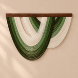 A wall-mounted decorative hanging made of layered curved strands in dark and light green with off-white sections, suspended from a dark brown wooden bar against a warm beige wall.
