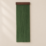 A vertical green fringe wall hanging with a dark brown wooden top bar, displayed against a warm beige wall with soft shadow overlays.