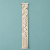 A vertical cream macrame wall hanging with geometric woven patterns and fringe, suspended from a wooden dowel against a solid teal-blue background.