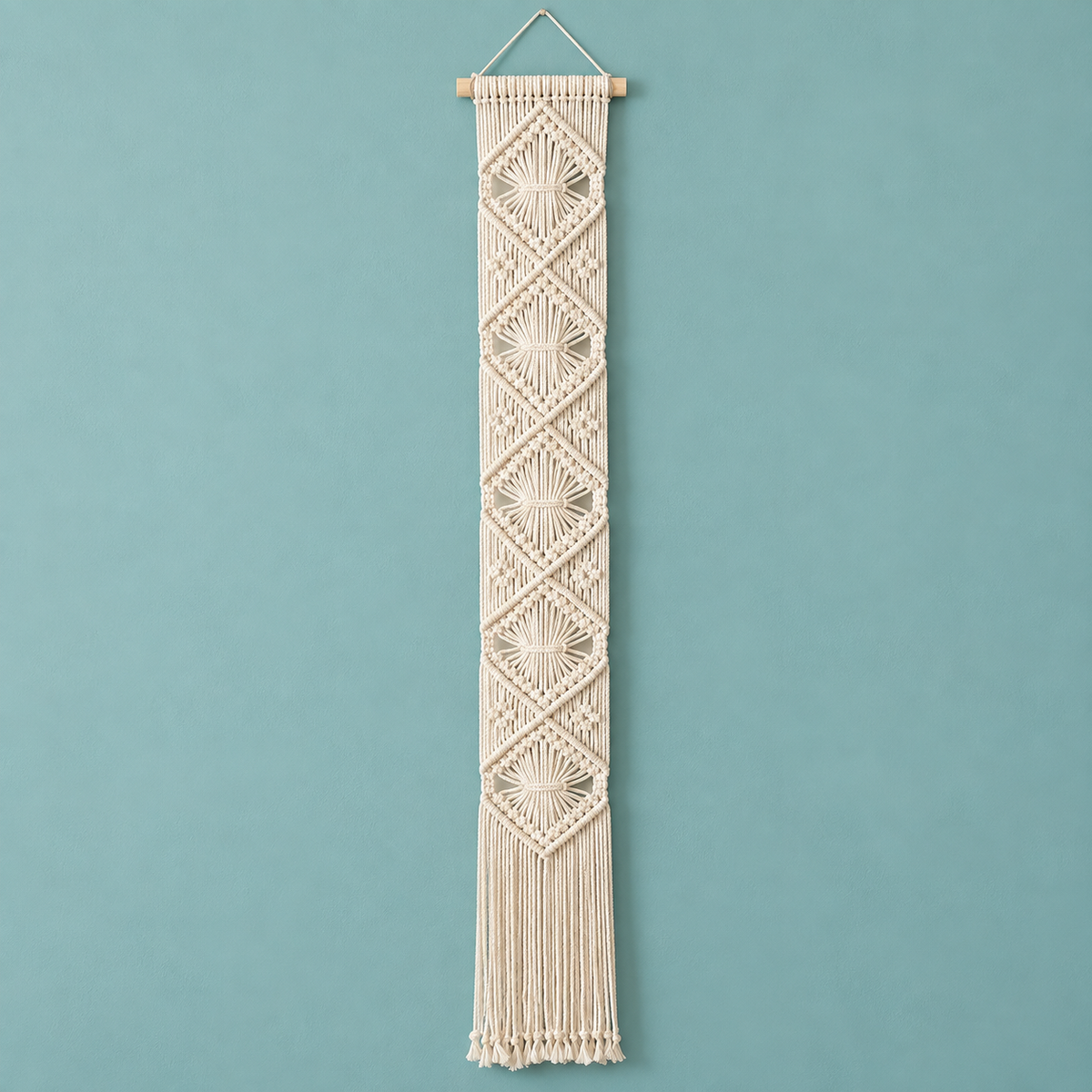 A vertical cream macrame wall hanging with geometric woven patterns and fringe, suspended from a wooden dowel against a solid teal-blue background.