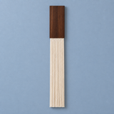A minimalist wall-mounted decorative piece featuring a dark brown wood block at the top and a long, narrow fringed section below in a light natural wood tone, set against a solid blue background.
