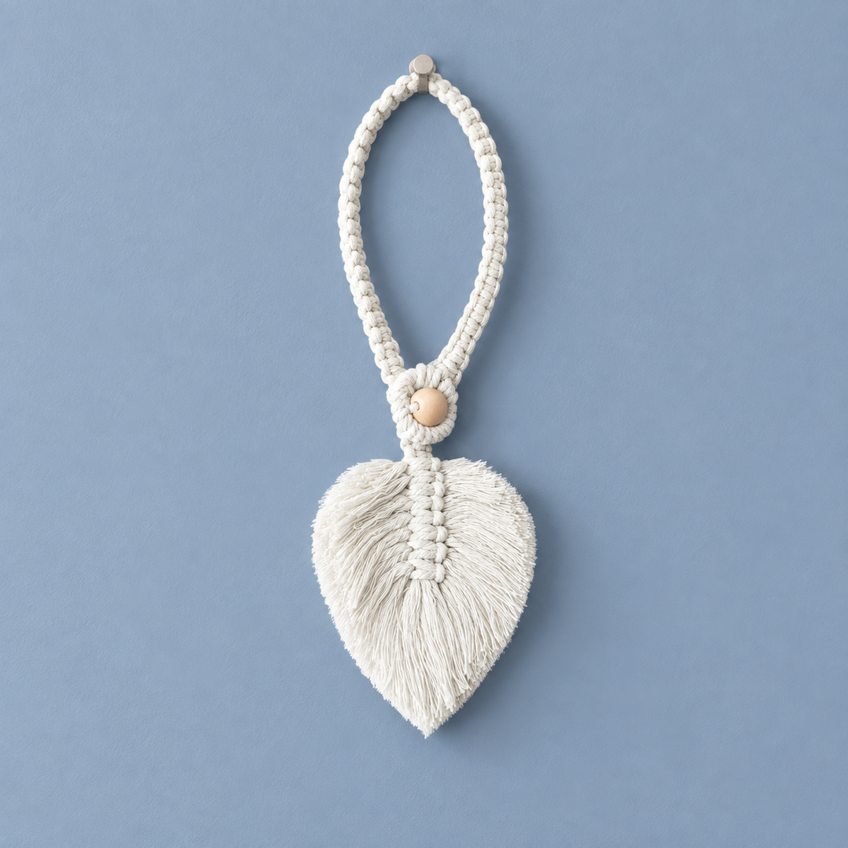 A cream-colored macrame leaf ornament with a tassel fringe and a small wooden bead accent, hanging against a solid dusty blue background.