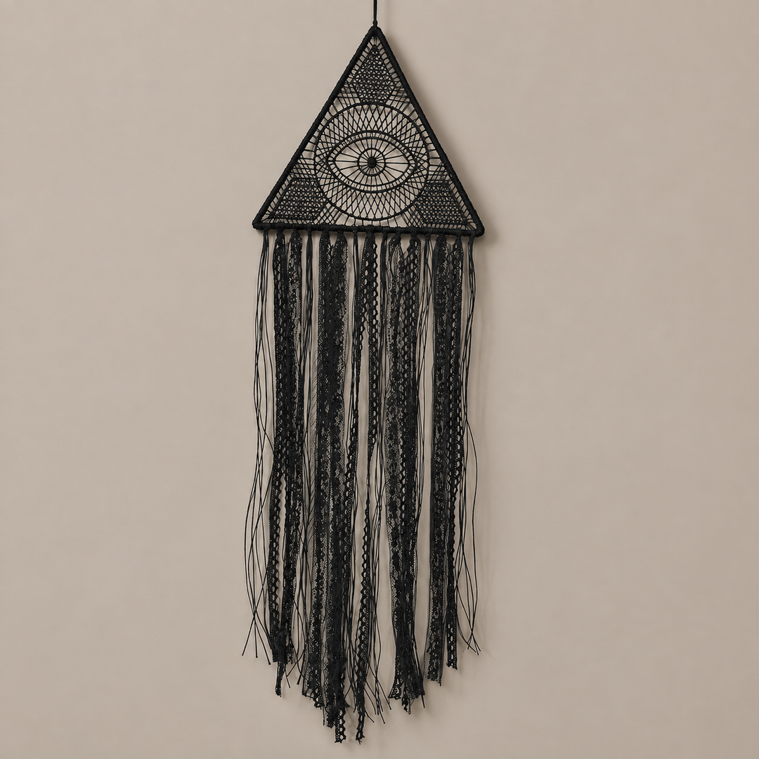 A black macrame wall hanging featuring a triangular top with an eye motif and long fringe strands, suspended against a plain beige wall.