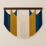 A bohemian-style macrame wall hanging with a dark wood bar, featuring a central cream fringe panel flanked by mustard yellow and deep teal fringe panels, mounted on a light beige wall.
