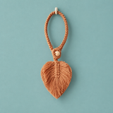A terracotta-colored braided keychain with a textured leaf-shaped tassel and a small wooden bead, hanging from a white wall hook against a solid teal background.